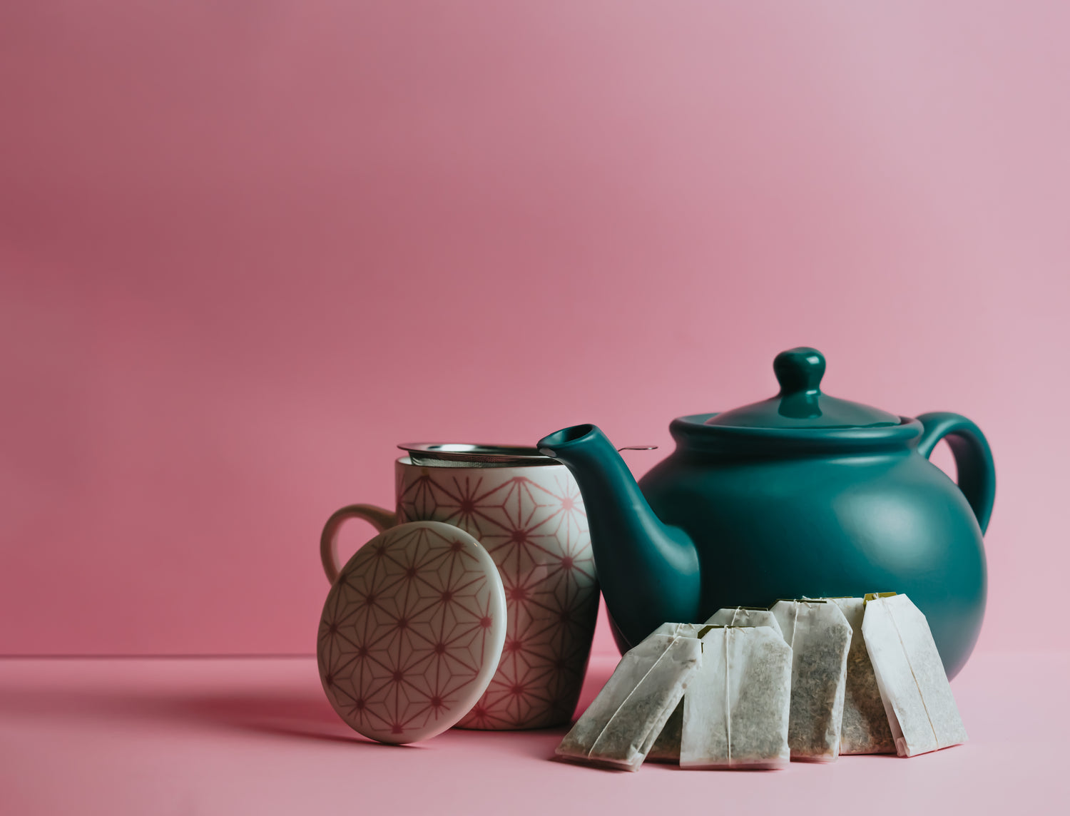 On a plain pink backgrounds sits a green ceramic teapot alongside a cup, and 5 teabags.