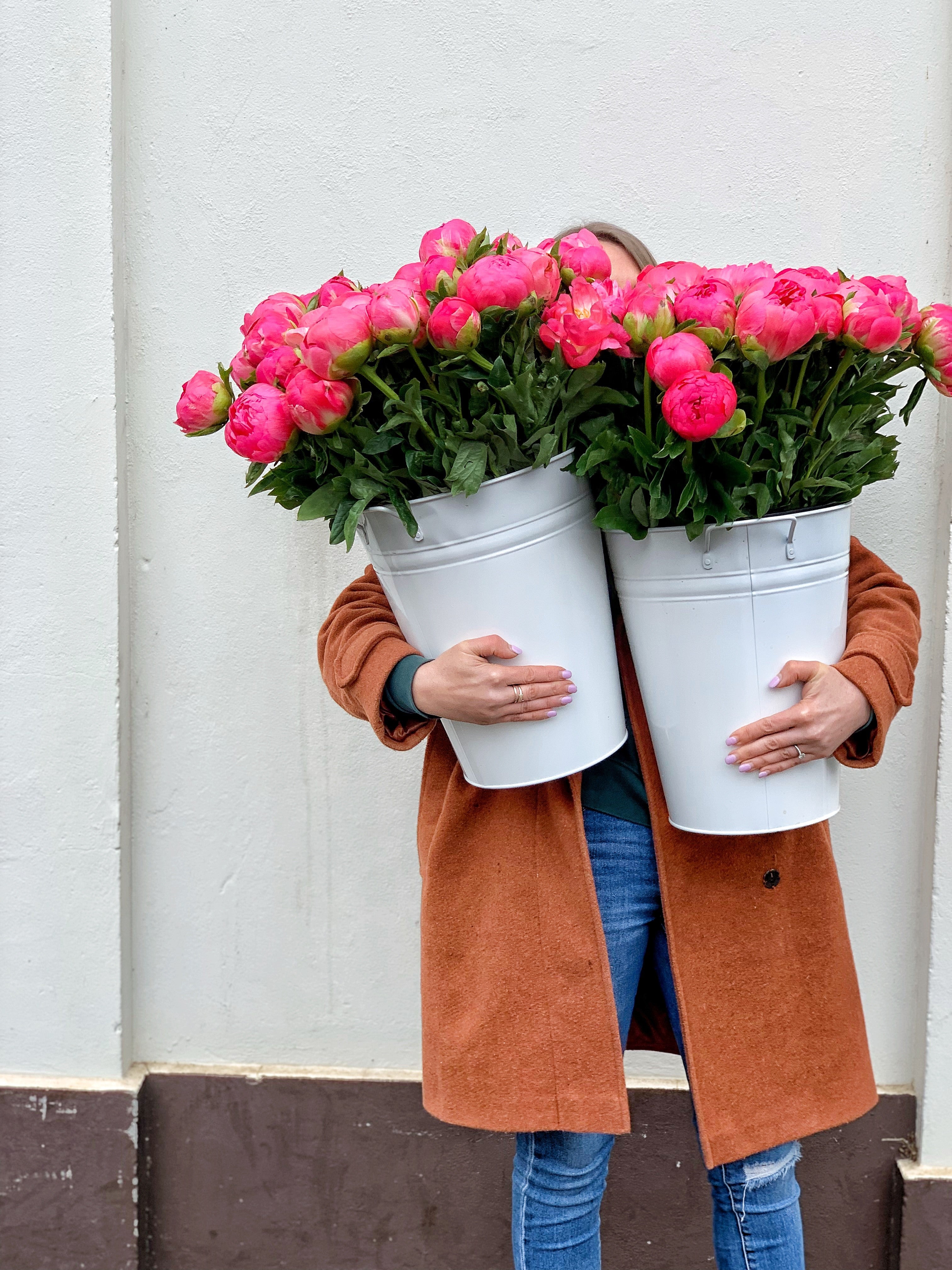 Lady holding two large white metal buckets that are full of deep pink peonies, so big we can't see her face